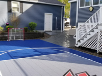 View from hoop end of small blue and grey basketball court with custom red H logo in Braintree, MA, showing customer's hockey net and patio they had installed to go with court.