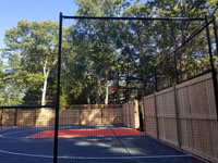 Looking through mesh side fence toward the hoop and key area of graphite and orange residential basketball court in Walpole, MA. Part of the court is fenced with a custom combination of cedar and mesh sections.