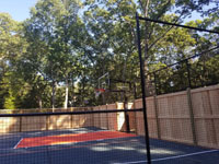Looking through mesh court fence at hoop end of graphite and orange home basketball court in Walpole, MA, including custom fencing that combines cedar fence with a topper of mesh.