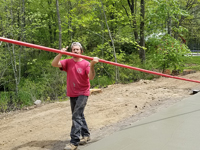 Worker uses long-handled tool for smoothing wet cement for base to construct black and grey home backyard basketball court in Wellesley, MA.