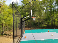 Dover, MA residential basketball court in green and silver with partial fencing, light, hoop system, and net accessories to add sports beyond basketball. Four balls the owner placed around the key feature in this shot.
