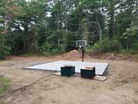 Green and red residential basketball court in Middleborough, MA. Shown here: Completed concrete base and installed goal system, with pallets of court tyles awaiting installation.