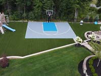 Low, angled drone view highlighting place of honor of silver and blue backyard basketball court in a beautifully updated landscape in Middleton, MA.