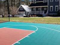 Residential basketball court in Norwell, MA, including goal system, mesh fencing, and an emerald green and rust red tile sport surface. Some trees and stumps were removed to make room in the yard and provide easy work access.