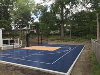 Angled end view of a shaded backyard basketball court in Randolph, MA, with primary tiles in a color that isn't always sure whether it wants to be blue or black .