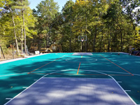 View of most of large emerald green and titanium backyard basketball court in Bolton, MA, still under last of construction. Besides full-featured basketball court and hoops, plus containment fencing, middle of court is lined for pickleball, to be played with a portable net not shown.